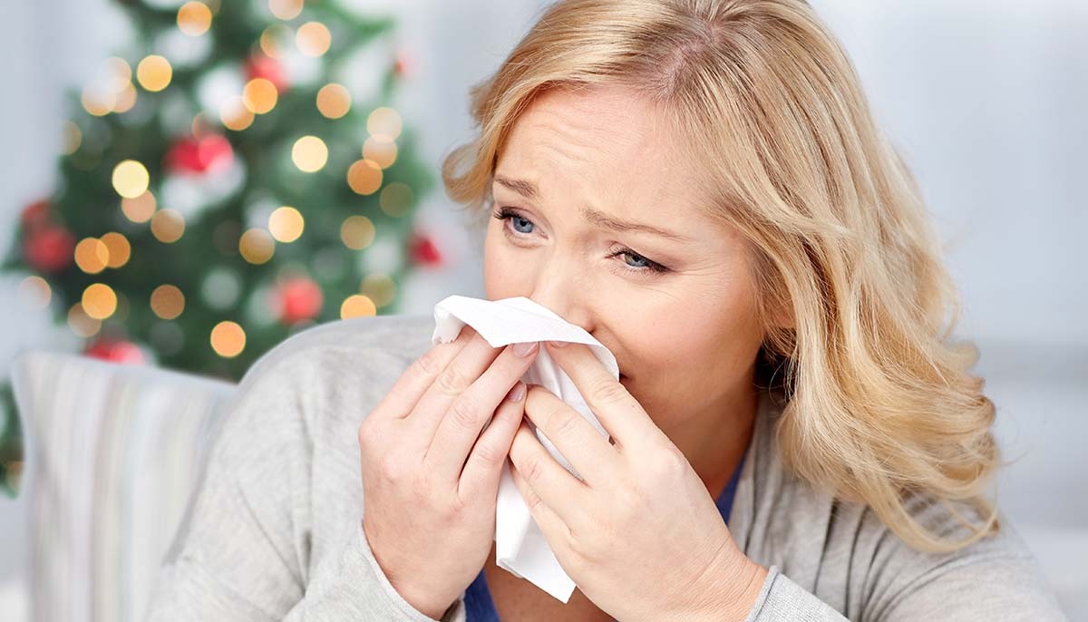 a blonde woman blows her nose with a christmas tree in the background