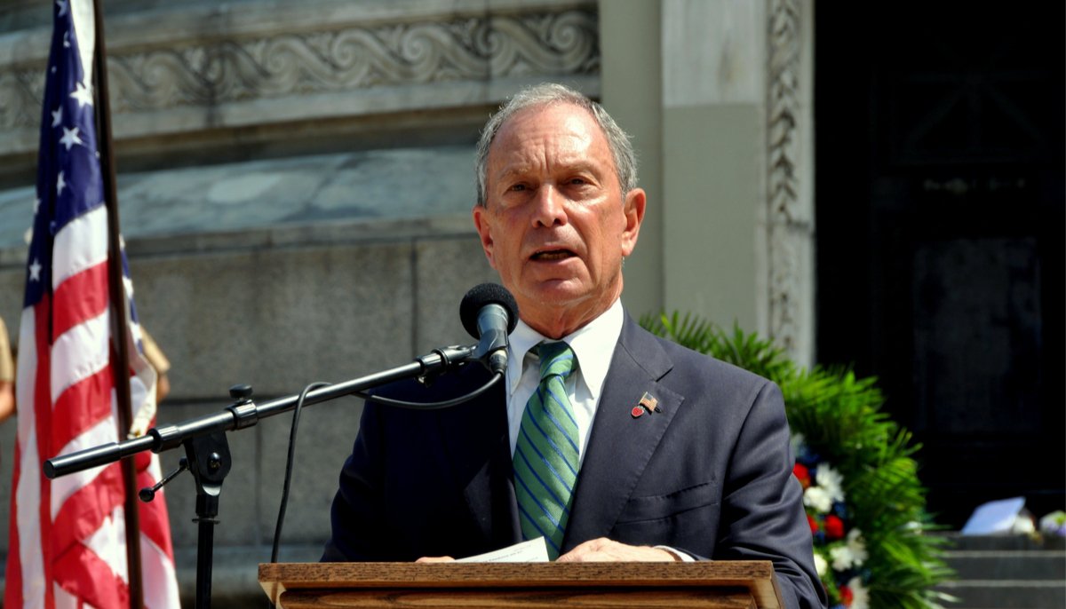 michael bloomberg speaking into a podium mic outside of a building, with a flag next to him