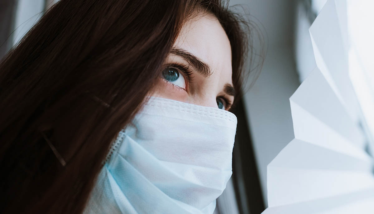  woman wearing medical mask looks out of window blinds