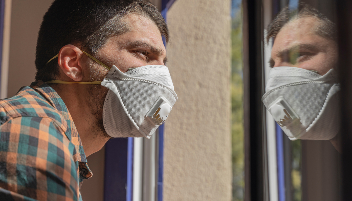 A man wearing a surgical face mask looks out his window