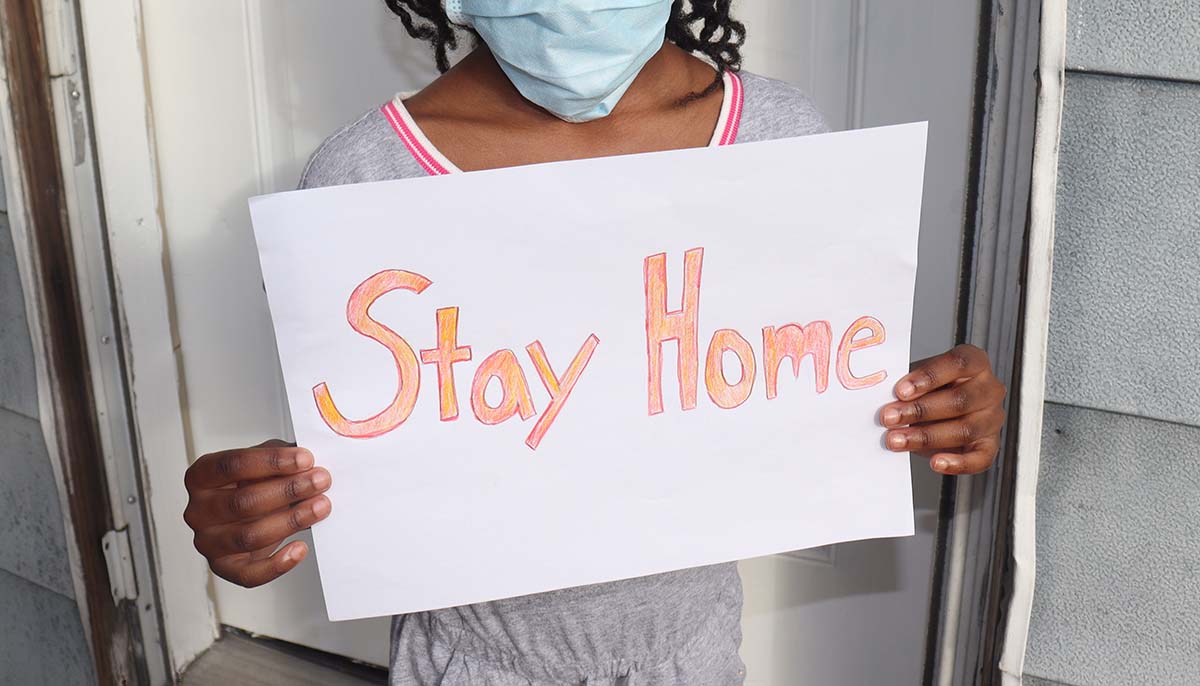 a child wears a mask while carrying a sign that reads stay home