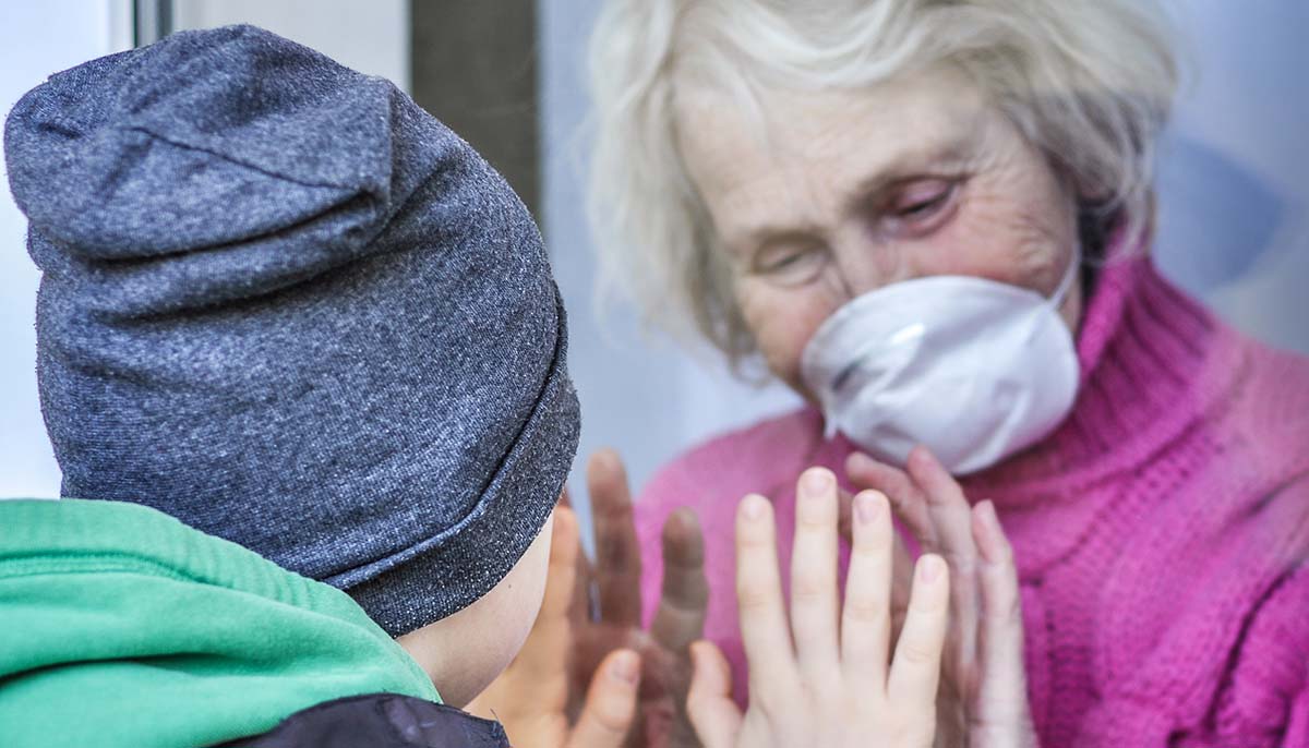 grandmother wearing face mask plays with grandson through a window
